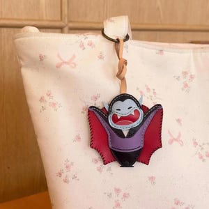 May include: A cream-colored tote bag with a pink floral and ribbon pattern features a leather vampire bat keychain. The bat has a light blue face, red mouth with white fangs, a purple chest, and dark red wings, attached by a tan leather strap.