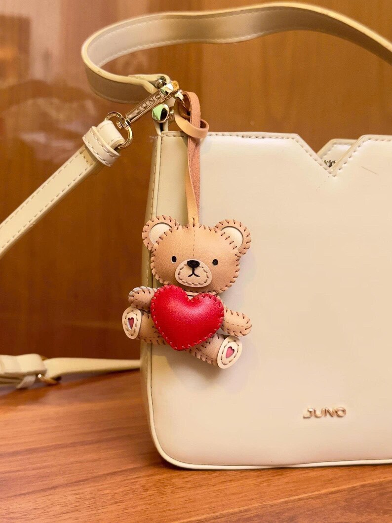 May include: A cream-colored handbag with a matching shoulder strap. A small, brown teddy bear charm with a red heart is attached to the bag. The bag has the word "JUNO" printed on the front.