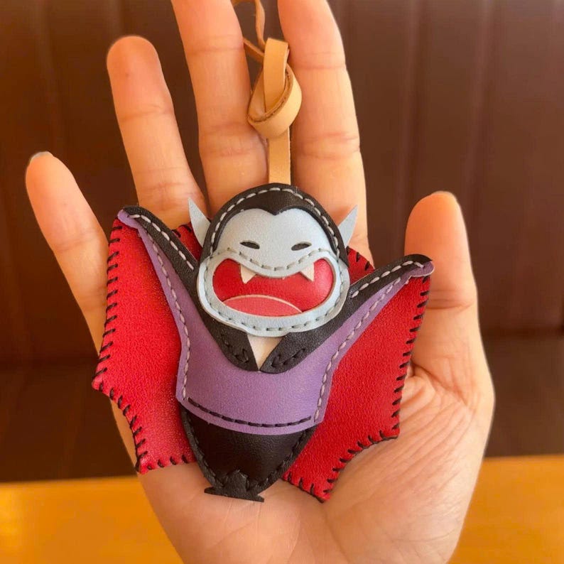 May include: A handmade leather keychain in the shape of a cartoon vampire. The vampire has a light blue face, a purple vest, and red wings. The keychain has a leather loop for attachment.