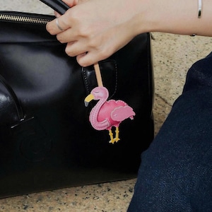 May include: A pink flamingo bag charm featuring a yellow beak with a grey tip and yellow legs, attached to a light-colored wooden stick. The flamingo has stitched detailing along its body and wings, and it is suspended against a black handbag.