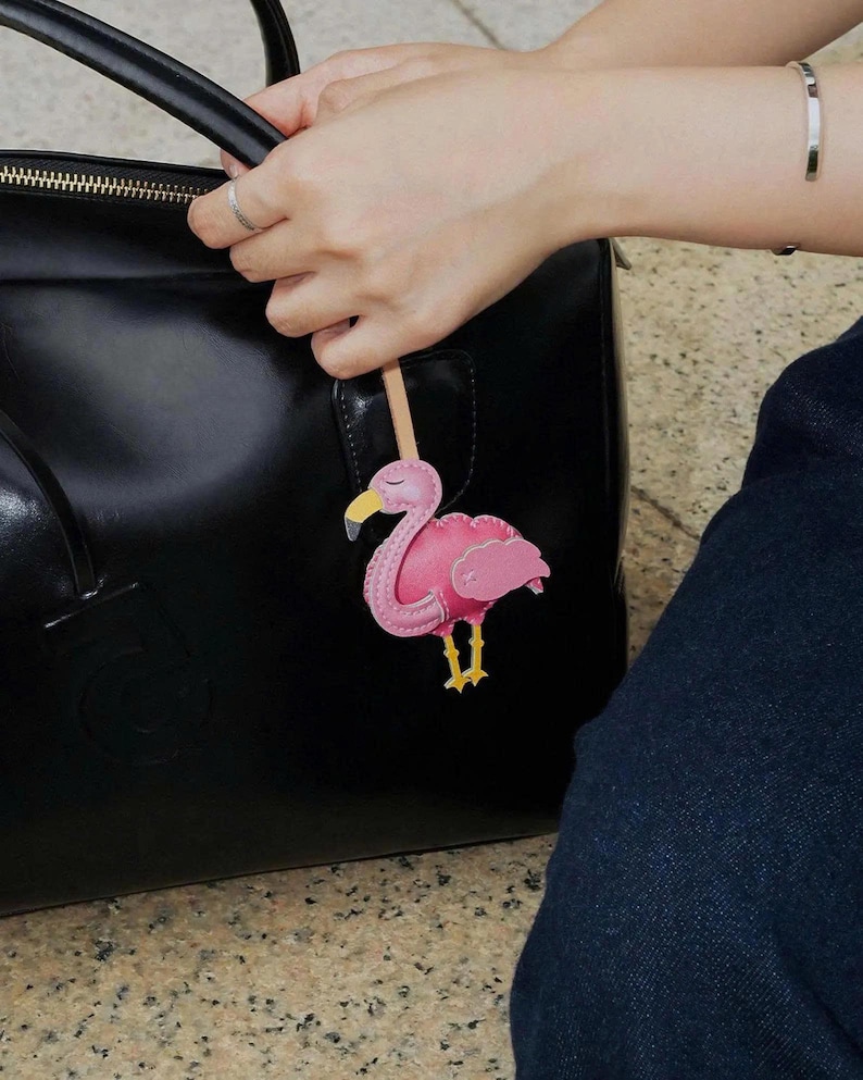 May include: A pink flamingo bag charm featuring a yellow beak with a grey tip and yellow legs, attached to a light-colored wooden stick. The flamingo has stitched detailing along its body and wings, and it is suspended against a black handbag.
