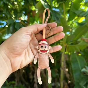 May include: A handmade sock monkey keychain. The keychain features a light brown monkey with white accents on the hands and feet. The monkey has a red hat and a stitched mouth. The keychain is attached to a leather strap and a metal ring.