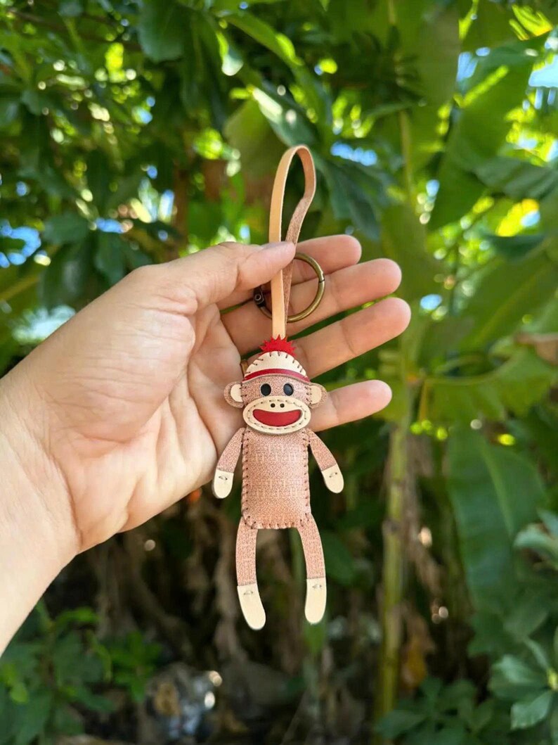 May include: A handmade sock monkey keychain. The keychain features a light brown monkey with white accents on the hands and feet. The monkey has a red hat and a stitched mouth. The keychain is attached to a leather strap and a metal ring.