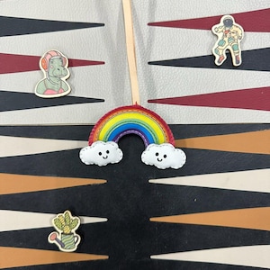 May include: A rainbow-colored felt rainbow with a smiling face and two white clouds with smiling faces. The rainbow is attached to a wooden stick. The rainbow is on a black and white patterned surface.