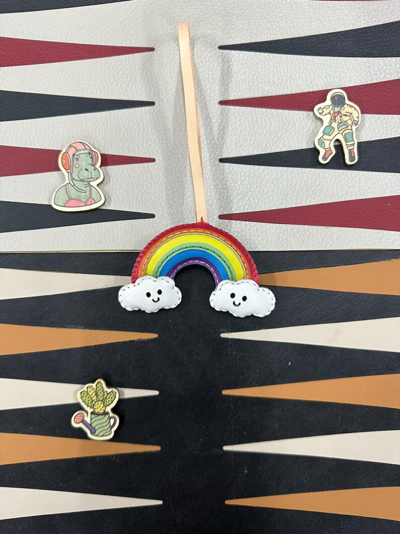 May include: A rainbow-colored felt rainbow with a smiling face and two white clouds with smiling faces. The rainbow is attached to a wooden stick. The rainbow is on a black and white patterned surface.