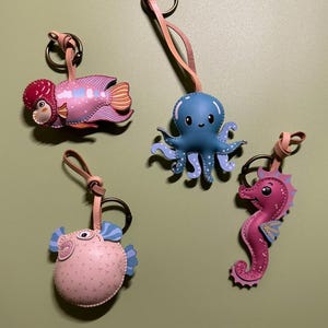 Può includere: Quattro portachiavi in pelle a forma di creature marine: un pesce rosa a righe, un polpo blu con pois bianchi, un pesce palla rotondo rosa chiaro con pinne blu e un cavalluccio marino rosa scuro. Ogni portachiavi ha un anello in metallo e un cinturino.