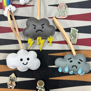 May include: Four felt cloud ornaments with different expressions. A gray cloud with an angry face and lightning bolts, a white cloud with a happy face, a gray cloud with a sad face and raindrops, and a rainbow with a white cloud.