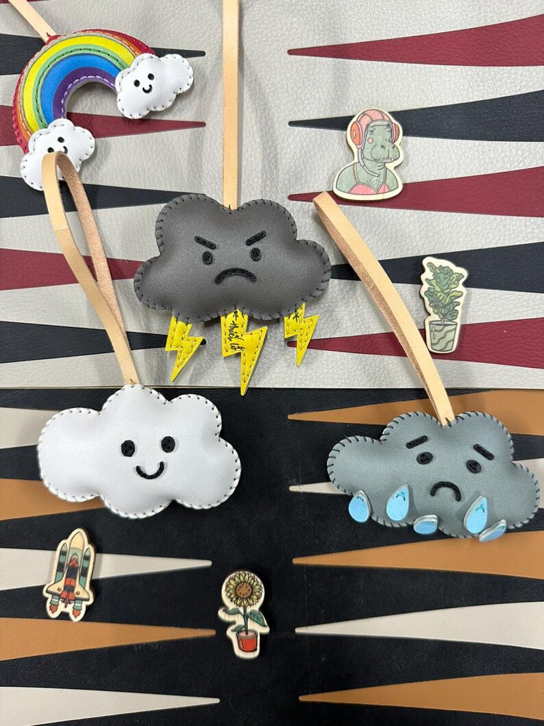 May include: Four felt cloud ornaments with different expressions. A gray cloud with an angry face and lightning bolts, a white cloud with a happy face, a gray cloud with a sad face and raindrops, and a rainbow with a white cloud.