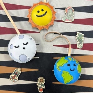 May include: A set of four leather ornaments shaped like the sun, moon, earth, and a rocket ship. Each ornament has a smiling face and is decorated with stitching. The ornaments are hanging from a white background with a black and red striped pattern.