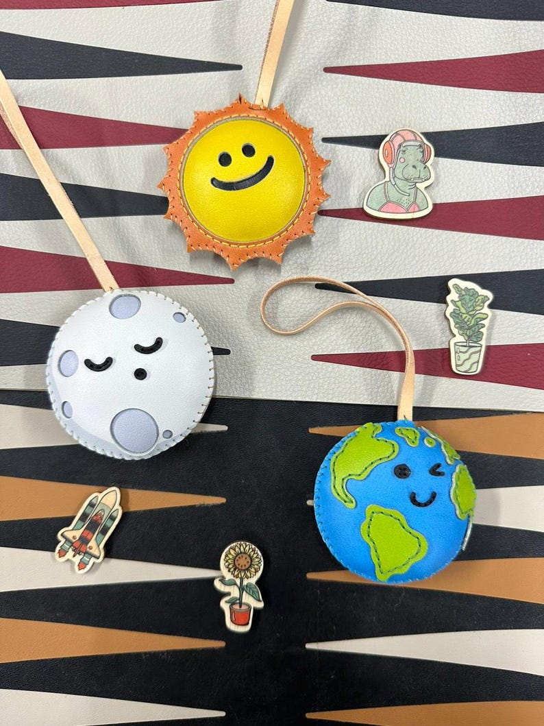 May include: A set of four leather ornaments shaped like the sun, moon, earth, and a rocket ship. Each ornament has a smiling face and is decorated with stitching. The ornaments are hanging from a white background with a black and red striped pattern.