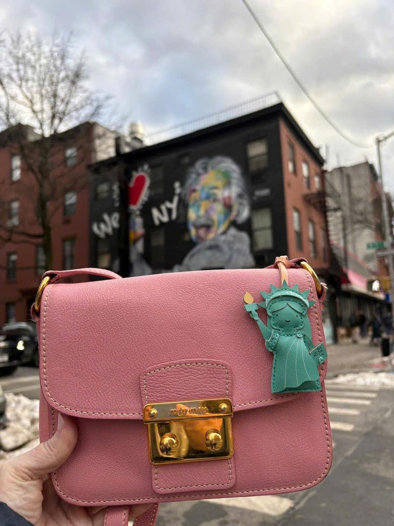 May include: A pink leather crossbody bag with a gold clasp and hardware. A green Statue of Liberty charm hangs from the strap. The background shows a street scene with a mural that says "We NY".