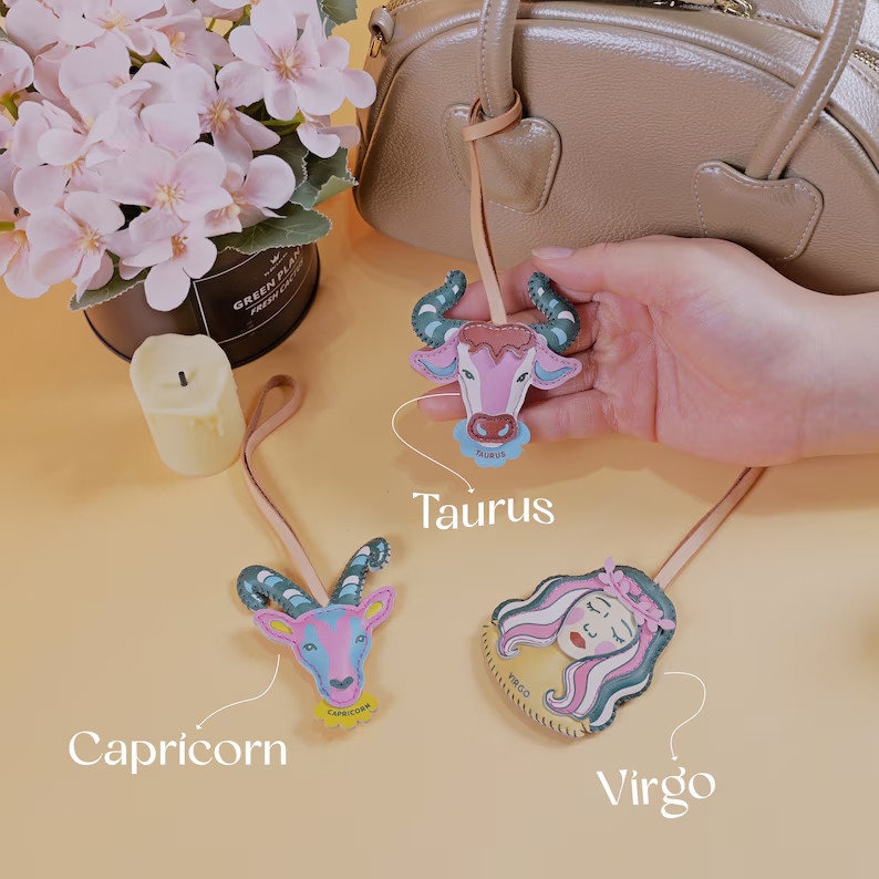 Puede incluir: Tres dijes del zodiaco de cuero con un esquema de color rosa, verde y azul. Los dijes tienen forma de toro, cabra y rostro de mujer. Los dijes est&aacute;n etiquetados como "Tauro", "Capricornio" y "Virgo".