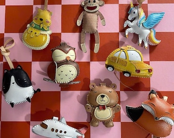 May include: A selection of leather ornaments in diverse shapes, such as a cat, owl, bear, fox, aeroplane, taxi, sock monkey, unicorn, and a yellow character. Each ornament has a leather loop for hanging, set against a pink and red checkered background.