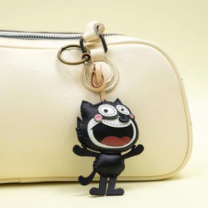 May include: A black and white cartoon cat keychain featuring a wide grin, large eyes, and pink cheeks, attached to a brass-colored metal ring. The keychain is suspended from the handle of a smooth, cream-colored handbag with visible stitching.