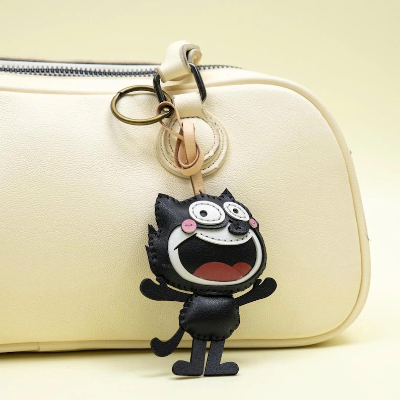 May include: A black and white cartoon cat keychain featuring a wide grin, large eyes, and pink cheeks, attached to a brass-colored metal ring. The keychain is suspended from the handle of a smooth, cream-colored handbag with visible stitching.