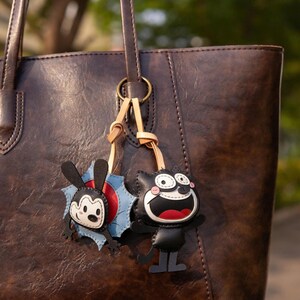 May include: A brown leather tote bag with two cartoon character keyrings attached. One keyring is a black cat with a red mouth and white eyes. The other is a black and white bunny with a blue and red background.