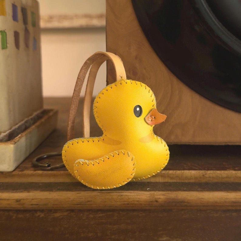 May include: A yellow leather duck keychain with a brown leather strap. The duck has a stitched outline, a small orange beak, and a black eye. The keychain is on a wooden surface.