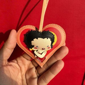 May include: A heart-shaped leather ornament featuring a cartoon character with black hair and a red dress. The ornament has a yellow and orange heart design, with a beige ribbon for hanging. The background is a solid red.
