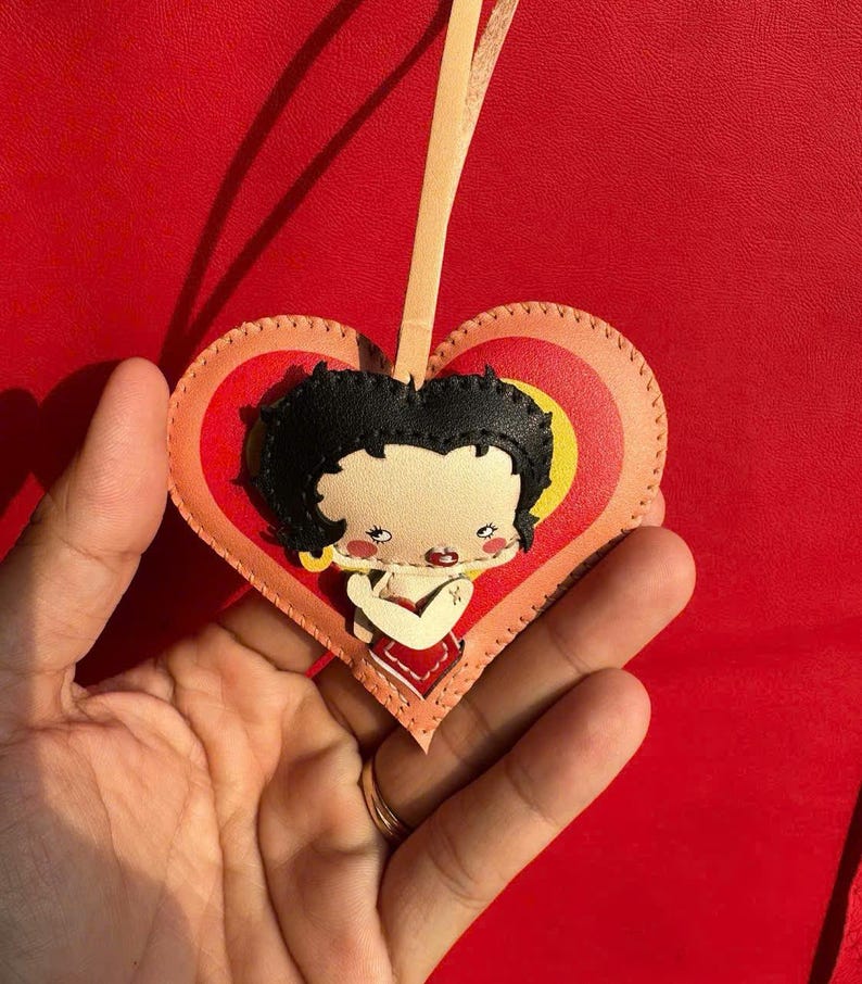 May include: A heart-shaped leather ornament featuring a cartoon character with black hair and a red dress. The ornament has a yellow and orange heart design, with a beige ribbon for hanging. The background is a solid red.