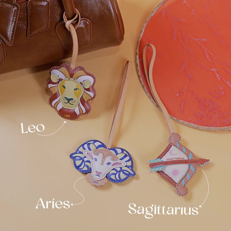 Puede incluir: Tres llaveros de cuero con signos del zod&iacute;aco: un le&oacute;n para Leo, un carnero para Aries y una flecha para Sagitario. Los llaveros est&aacute;n sobre un fondo amarillo claro.