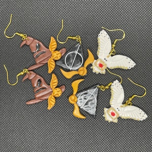 May include: A set of six Harry Potter themed earrings. The earrings feature a Sorting Hat, a Golden Snitch, a Deathly Hallows symbol, and a Snowy Owl. The earrings are made of polymer clay and have gold hooks.