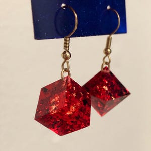 May include: A pair of dangle earrings featuring red, geometric, cube-shaped pendants with gold flecks. The earrings have gold-colored hooks and are displayed against a blue background.