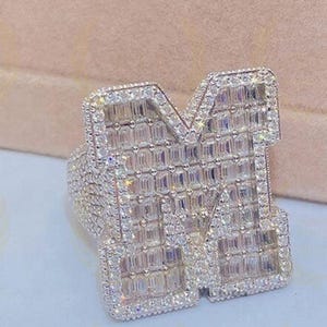 Könnte beinhalten: Ein funkelnder, silberfarbener Ring in Form des Buchstabens "M", besetzt mit zahlreichen kleinen, glitzernden Steinen. Der Ring hat ein aufwendiges Design und wird vor einem weichen, neutralen Hintergrund präsentiert, der seinen luxuriösen Look unterstreicht.