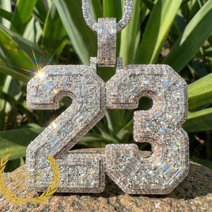 Könnte beinhalten: Ein silberner Anhänger in Form der Zahl 23, besetzt mit zahlreichen Diamanten. Das Design des Anhängers weist eine kontrastierende Metallkontur auf. Die Diamanten sind rechteckig geschliffen. Der Anhänger hängt an einer passenden Kette.