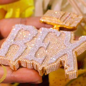 Könnte beinhalten: Ein individueller Anhänger mit dem Wort "RAY" in einem funkelnden, diamantähnlichen Material. Der Anhänger hat einen goldfarbenen Rand und wird in einer Hand gehalten. Der Hintergrund ist verschwommen mit gelben und weißen Farben.