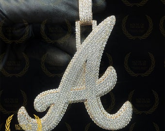May include: A large, gold-tone pendant in the shape of the letter 'A' is completely covered in small, clear, sparkling stones. The pendant is on a black background.