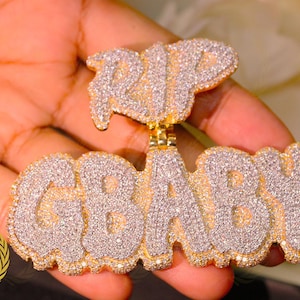 Könnte beinhalten: Goldener Anhänger mit den Wörtern "RIP" und "GBABY", besetzt mit glitzernden weißen Steinen. Der Anhänger wird in einer Hand gehalten, was die feinen Details und die Handwerkskunst des Schmucks zeigt.