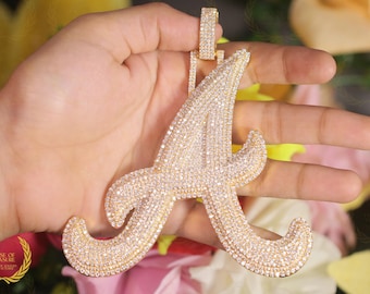 May include: A gold-colored pendant in the shape of the letter "A", encrusted with numerous small, sparkling stones. The pendant is held up by a small loop at the top. The background is blurred, with hints of yellow and white flowers.