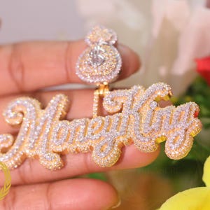 Könnte beinhalten: Goldfarbener Anhänger mit dem Schriftzug "Money King" in Schreibschrift, besetzt mit kleinen, funkelnden Steinen. Der Anhänger hat auch ein Geldsack-Design oben, ebenfalls mit Steinen verziert. Der Schmuck wird von einer goldenen Kette gehalten.