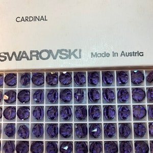 Può includere: Una vista ravvicinata di una scatola di cristalli Swarovski Cardinal da 10 mm. La scatola è bianca con testo nero e contiene 1 grosso di cristalli. I cristalli sono di colore viola scuro e sono disposti in uno schema a griglia.