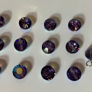 Puede incluir: Un grupo de 13 cuentas redondas de vidrio morado facetadas con brillo iridiscente. Una cuenta está unida a un alambre de plata.