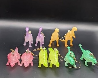 Könnte beinhalten: Ohrringe in Dinosaurierform in verschiedenen Farben: Rosa, Grün, Gelb und Lila. Die Ohrringe bestehen aus einem durchscheinenden Material und haben goldfarbene Haken. Die Dinosaurier sind in verschiedenen Posen dargestellt.