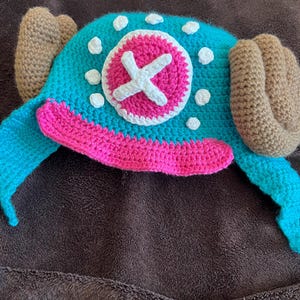 May include: A handmade crochet hat in teal, pink, and brown. The hat features a pink and white circular design with a white cross in the center, and brown ear-like appendages on each side. White polka dots are scattered across the teal surface.