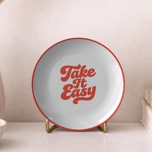 Puede incluir: Un plato blanco con borde rojo y la frase "Take It Easy" en rojo. El plato se exhibe sobre un soporte dorado. Un jarr&oacute;n blanco con plantas secas y un cuenco con nueces en el fondo.