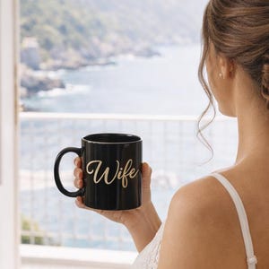 Puede incluir: Una taza de café negra con la palabra "Wife" en escritura dorada. La taza la sostiene una persona con un vestido blanco, sentada cerca de una ventana con vistas a una pintoresca vista costera.