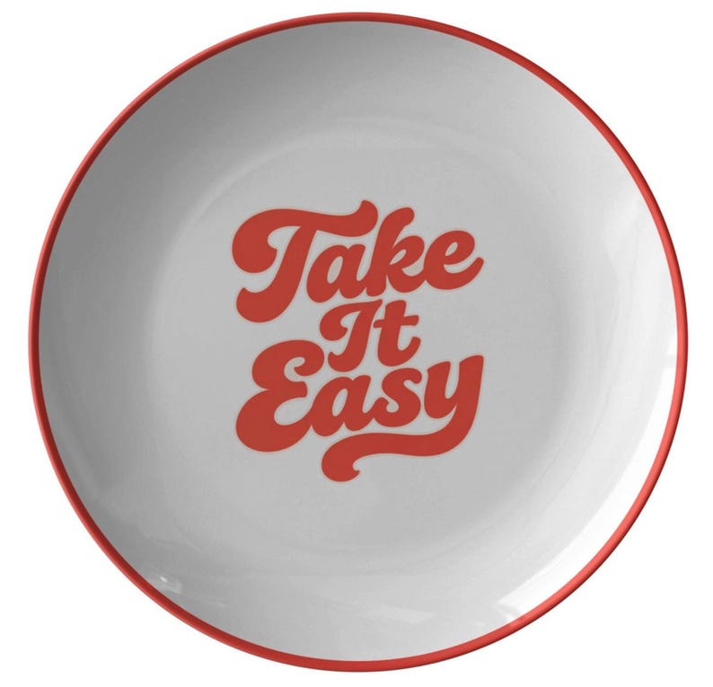 Puede incluir: Plato de cer&aacute;mica blanco con borde rojo. El plato presenta la frase "Take It Easy" en una fuente retro roja. El dise&ntilde;o es simple y limpio, adecuado para el uso diario.