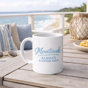 Puede incluir: Taza de cerámica blanca con el texto "Montauk ALWAYS A GOOD IDEA" en escritura azul claro. La taza está sobre una mesa de madera con una escena de playa al fondo.