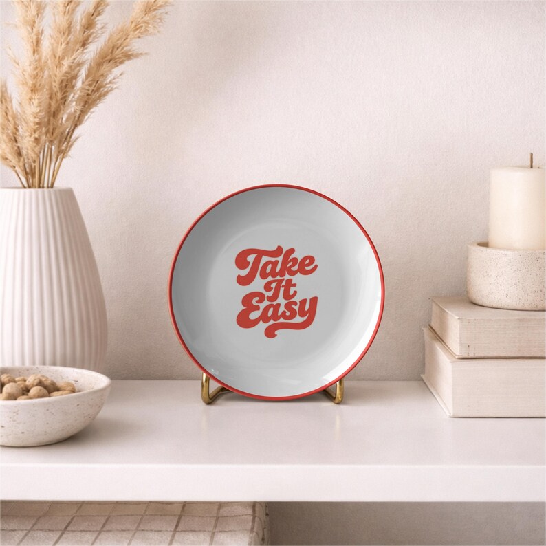 Puede incluir: Un plato blanco con borde rojo y la frase "Take It Easy" en rojo. El plato se exhibe sobre un soporte dorado. Un jarr&oacute;n blanco con plantas secas y un cuenco con nueces en el fondo.