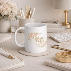 Puede incluir: Taza de cerámica blanca con la inscripción dorada "Boss Mode", rodeada de ilustraciones florales melocotón y crema. La taza está sobre un posavasos beige, con un bolígrafo y un cuaderno en primer plano.