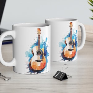 May include: Two white ceramic mugs with a watercolor design of an acoustic guitar. The guitar is brown with a light wood grain and has blue and white watercolor splatters behind it.