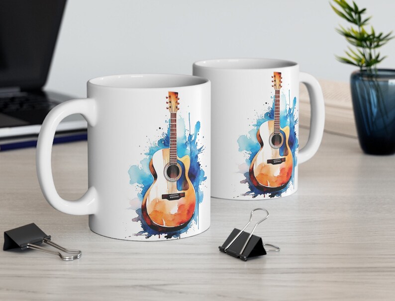 May include: Two white ceramic mugs with a watercolor design of an acoustic guitar. The guitar is brown with a light wood grain and has blue and white watercolor splatters behind it.