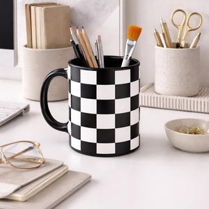 Puede incluir: Una taza a cuadros blancos y negros llena de lápices, bolígrafos y un pincel. La taza está sobre un escritorio blanco junto a un cuenco pequeño, cuadernos y un par de gafas. Otros recipientes con material de oficina están en el fondo.