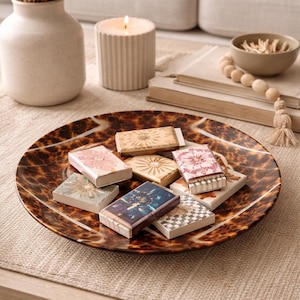 May include: A decorative brown and tan tortoiseshell-patterned plate holds several small matchboxes with floral and geometric designs. A lit candle and a vase with dried plants are in the background, creating a cozy, home decor scene.