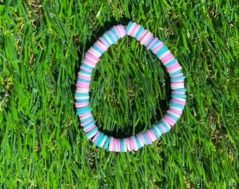 May include: A pink and teal beaded bracelet with a circle shape. The bracelet is made of small, round beads that are strung together on a string. The beads are arranged in a pattern of pink and teal.