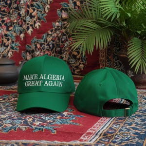 Op de afbeelding: Twee groene baseballpetten. Een pet heeft de tekst "Make Algeria Great Again" in wit geborduurd op de voorkant. De andere pet is effen.
