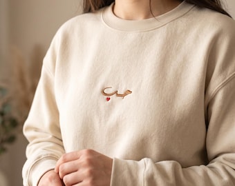 May include: A beige crewneck sweatshirt featuring a small, centered embroidery of the Arabic word for love in a brown thread, accented with a tiny red heart positioned directly beneath the text on the upper chest area of the garment.